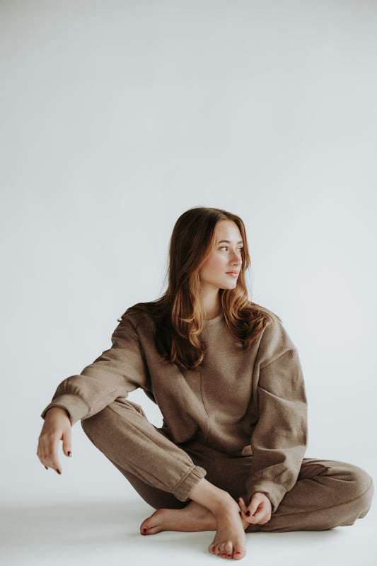 A woman is sitting on the ground, looking on the left and wearing the classic fleece set in the color chocolate brown