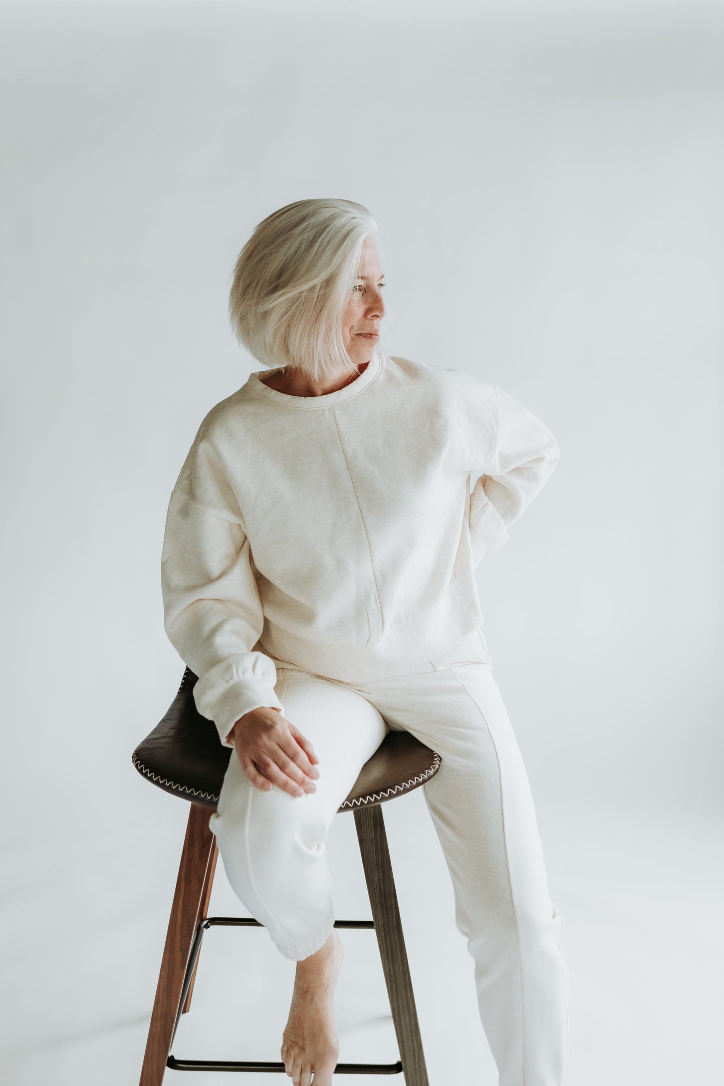 A woman is sitting on a chair and wearing the classic fleece crewneck and sweatpants in the color beige 