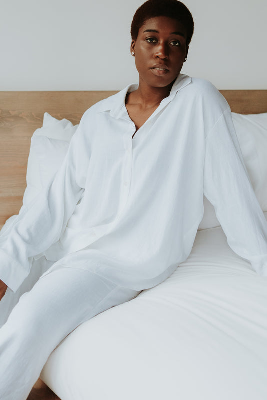 a woman is sitting on a bed and wearing the staple linen blouse and pants in white